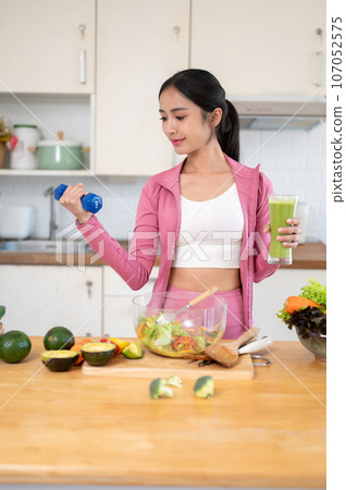 A slim Asian woman in sportswear is holding a dumbbell and a glass of green smoothie in the kitchen. 107052575