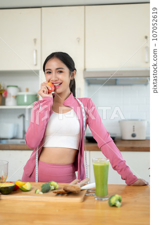 A beautiful and healthy Asian woman in sportswear stands at a kitchen table in the kitchen. A beautiful and healthy Asian woman in sportswear stands at a kitchen table in the kitchen. 107052619