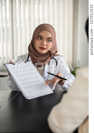 An Asian Muslim female doctor shows information on a clipboard to a patient during a medical checkup 107052835