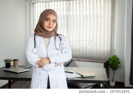 An attractive Asian Muslim female doctor in a uniform stands with her arms crossed in her office. 107052841
