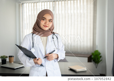 A professional Asian Muslim female doctor in a uniform and hijab stands in an office at a hospital. 107052842