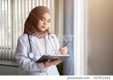 A focused Asian Muslim female doctor is checking her medical cases on a medical clipboard. A focused Asian Muslim female doctor is checking her medical cases on a medical clipboard. 107052849