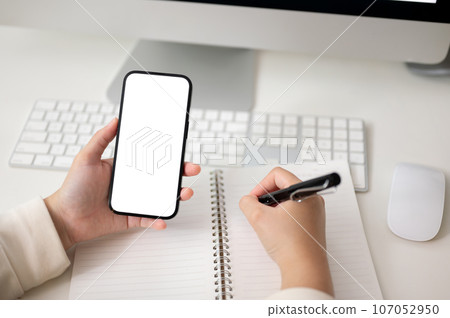A female using her smartphone and taking notes on her notebook at her computer desk. 107052950