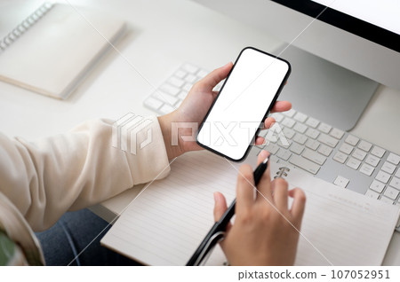A female using her smartphone and taking notes on her notebook at her computer desk. 107052951