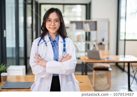 A professional Asian female doctor in a white gown stands with her arms crossed in her office 107053487