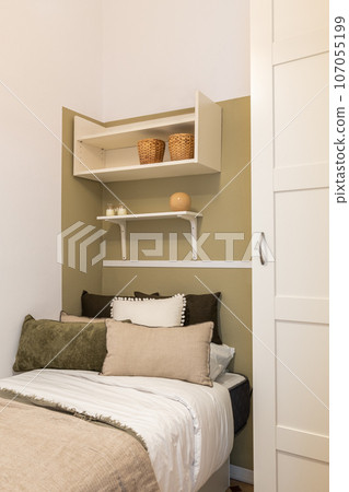 Vertical shot of a single bed with pillows and a duvet on olive white wallpaper and shelves and a wardrobe in a small hotel room. Business trip and travel concept Vertical shot of a single bed with pillows and a duvet on olive white wallpaper and shelves and a wardrobe in a small hotel room. Business trip and travel concept 107055199
