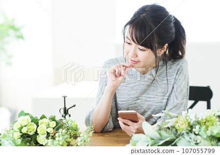 一位年輕女子在客廳一邊使用智慧型手機一邊插花 一位年輕女子在客廳一邊使用智慧型手機一邊插花 107055799