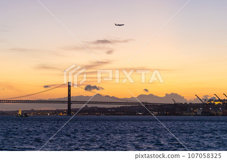 April 25 bridge over the Tagus River at sunset - the plane flies in the sky 107058325