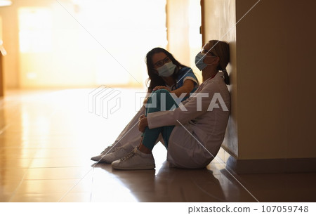 Two doctors in medical protective masks sit in corridor of medical facility 107059748