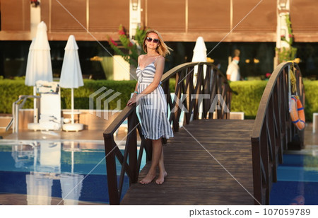 Woman in summer dress stands next to small bridge on hotel grounds 107059789