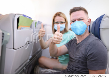 Man and woman in medical protective masks in aircraft cabin hold their thumbs up Man and woman in medical protective masks in aircraft cabin hold their thumbs up 107059797