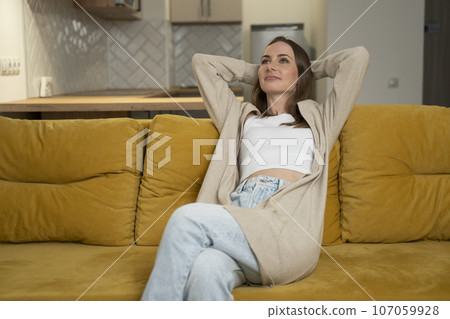 Calm brunette woman resting at home on a yellow sofa, with her eyes open and her hands behind her head 107059928