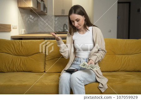 Woman with money and a calculator is sitting on a yellow sofa. A concerned woman with financial problems experiences stress when her rent or electricity costs are too high 107059929