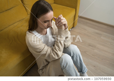Depressed woman is sitting on the floor in the living room near a yellow sofa 107059930