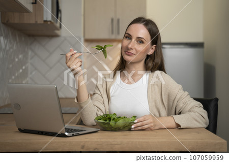 A woman is sitting at a table in the kitchen and enjoying a nutritious salad for lunch and using a laptop A woman is sitting at a table in the kitchen and enjoying a nutritious salad for lunch and using a laptop 107059959