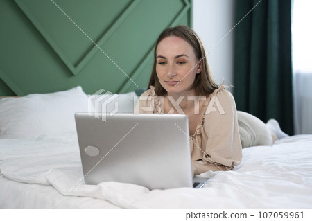 Beautiful young brunette woman working at home on a laptop lying on the bed 107059961