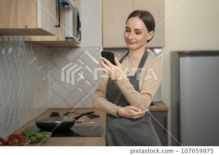 Young woman using a smartphone is studying at the kitchen table. A smiling woman reads a phone message. Brunette happy girl typing a text message Young woman using a smartphone is studying at the kitchen table. A smiling woman reads a phone message. Brunette happy girl typing a text message 107059975
