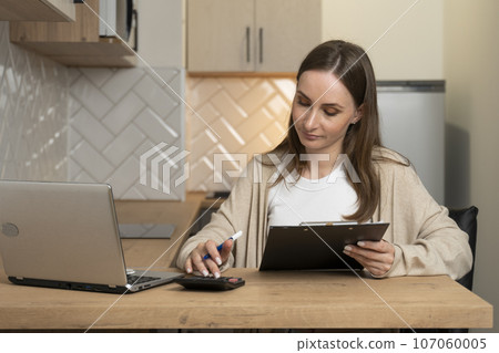 Serious young woman calculating finances, household expenses, working with project statistics using a laptop and a calculator, sitting at home in the kitchen Serious young woman calculating finances, household expenses, working with project statistics using a laptop and a calculator, sitting at home in the kitchen 107060005