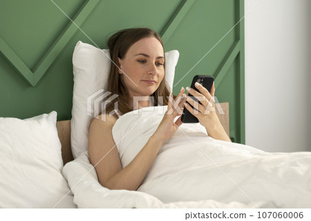 Portrait of a beautiful young brunette woman using a smart mobile phone on a bed with a white pillow-blanket in the bedroom interior 107060006