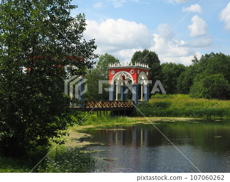 Spring landscape with fir trees and deciduous trees on the river bank with a beautiful wooden bridge, against a clear sky. Rural landscape, a beautiful place to relax. Spring landscape with fir trees and deciduous trees on the river bank with a beautiful wooden bridge, against a clear sky. Rural landscape, a beautiful place to relax. 107060262