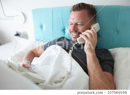 Smiling man lies in bed and talks on phone Smiling man lies in bed and talks on phone 107060599