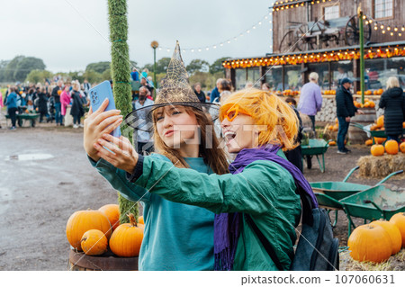 Two laughing female friends in fun glasses and witch hat having fun and making selfie on phone on decorated pumpkin farm. Selecting Thanksgiving and Halloween holidays decor. Autumn fall festive mood Two laughing female friends in fun glasses and witch hat having fun and making selfie on phone on decorated pumpkin farm. Selecting Thanksgiving and Halloween holidays decor. Autumn fall festive mood 107060631