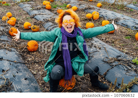 Emotional red head young woman in funny glasses with raised arms sitting on pumpkin on pumpkin patch field. Selecting best one for Thanksgiving and Halloween holidays. Autumn positive festive mood. Emotional red head young woman in funny glasses with raised arms sitting on pumpkin on pumpkin patch field. Selecting best one for Thanksgiving and Halloween holidays. Autumn positive festive mood. 107060632