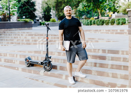 Handsome man holding laptop in his hand standing near electric scooter. Active lifestyle, backpacker traveler. Handsome man holding laptop in his hand standing near electric scooter. Active lifestyle, backpacker traveler. 107061976