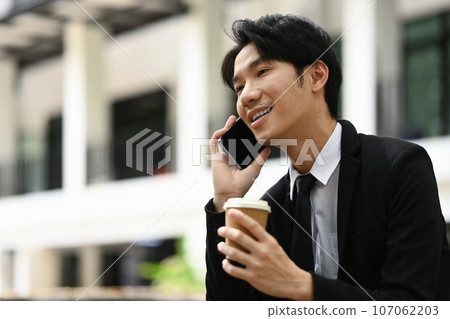 Positive asian businessman having pleasant phone conversation on mobile phone sitting outside office building 107062203