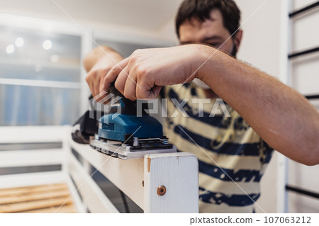 man rubs wooden child bed with power sander or grinder removing old paint 107063212