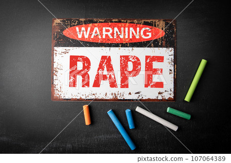 RAPE. Metal warning sign and colored pieces of chalk on a dark chalkboard background RAPE. Metal warning sign and colored pieces of chalk on a dark chalkboard background 107064389