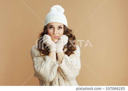 pensive trendy woman in beige sweater, mittens and hat on beige 107066285