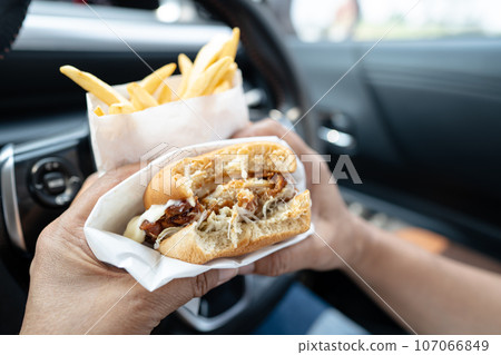 Asian lady holding hamburger and French fries to eat in car, dangerous and risk an accident. Asian lady holding hamburger and French fries to eat in car, dangerous and risk an accident. 107066849