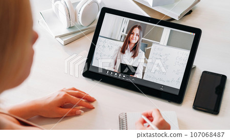 Online learning. Video lesson. Virtual chat. Distance education. Woman teacher on tablet screen performing presentation to female student writing notes at home workplace. 107068487