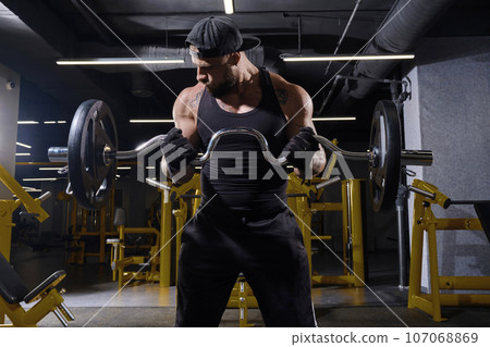 Tattooed male in black sport gloves, shorts, vest and cap. Lifting barbell, training his biceps, standing in dark gym with yellow equipment. Close up 107068869