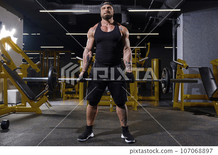 Bearded athlete in black sport gloves, shorts, vest, cap and sneakers. He lifting a barbell, posing in dark gym with yellow equipment. Full length 107068897
