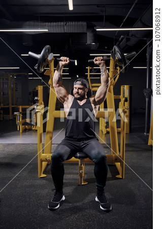 Tattooed, bearded male in black sweatpants, vest and cap. He performing a chest press while sitting on an exercise machine at dark gym. Full length Tattooed, bearded male in black sweatpants, vest and cap. He performing a chest press while sitting on an exercise machine at dark gym. Full length 107068912