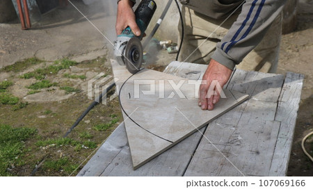 cutting large ceramic tiles with a grinder along a black oval outline in an outdoor tiler's workshop, trimming finishing tiles with a miter saw to the required size cutting large ceramic tiles with a grinder along a black oval outline in an outdoor tiler's workshop, trimming finishing tiles with a miter saw to the required size 107069166