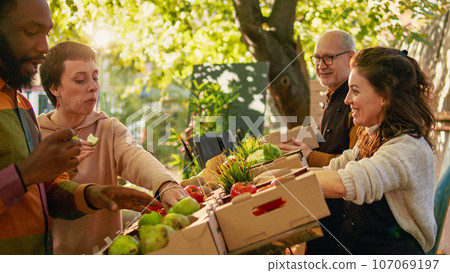 Diverse customers tasting slices of juicy apples before buying organic products at farmers market stand. Smiling couple doing produce sampling with fruits and veggies, laughing at marketplace. 107069197