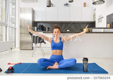 Young woman doing sports, meditating on yoga mat at home, holds hands sideways and breathing, relaxing mindful exercises 107069204
