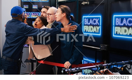 Security officer trying to calm people at store entrance during black friday sales rush, holding back shoppers obsessed with bargains. Guard working under pressure at shopping center. 107069223