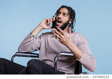 Emotional irritated arab man with disability talking on smartphone while sitting in wheelchair. Angry person with physical imparment making gestures while chatting on mobile phone Emotional irritated arab man with disability talking on smartphone while sitting in wheelchair. Angry person with physical imparment making gestures while chatting on mobile phone 107069225
