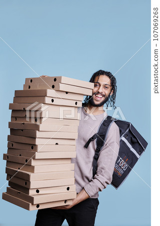 Carefree arab deliveryman carrying big pizza boxes pile and looking at camera with cheerful expression. Smiling restaurant delivery service courier standing huge stack of fast food packages 107069268