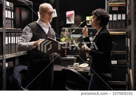Private detectives analyzing criminal investigations report, working overtime at solution to solve confidential crime in arhive room. Criminology department investigators checking evidence files Private detectives analyzing criminal investigations report, working overtime at solution to solve confidential crime in arhive room. Criminology department investigators checking evidence files 107069292