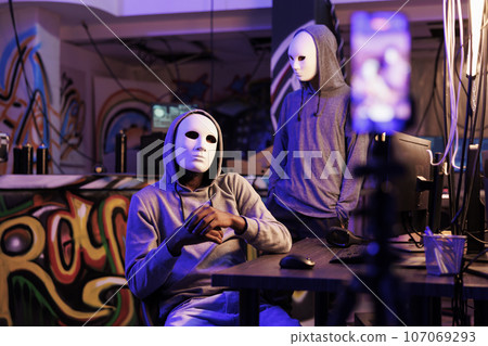 Scammers recording illegal activities on smartphone in abandoned warehouse. Anonymous internet thieves in masks engaging in cyberterrorism while broadcasting threat on dark web Scammers recording illegal activities on smartphone in abandoned warehouse. Anonymous internet thieves in masks engaging in cyberterrorism while broadcasting threat on dark web 107069293