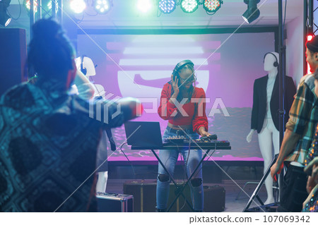 Woman musician using dj controller while performing on stage in nightclub. African american disc jokey playing and mixing electronic music during concert disco party in club 107069342