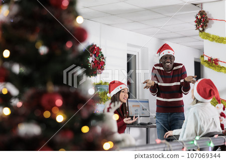 Diverse corporate team enjoying time spent together in festive decorated office during holiday season. Business personnel feeling Christmas spirit mood, having relaxing chat in xmas adorn workspace Diverse corporate team enjoying time spent together in festive decorated office during holiday season. Business personnel feeling Christmas spirit mood, having relaxing chat in xmas adorn workspace 107069344