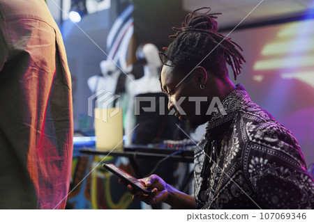 Young african american man using smartphone while resting in nightclub. Clubber scrolling social media and online pages on mobile phone while relaxing at discotheque party in club 107069346