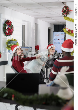 Colleagues having fun in xmas ornate office, making fun of each other at work during Christmas holiday season. Staff members laughing together in xmas adorn boardroom during coffee break 107069395