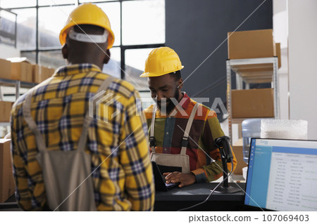 African american warehouse operators checking delivery schedule on laptop while standing at counter desk. Storehouse workers wearing overalls and helmets checking electronic orders list African american warehouse operators checking delivery schedule on laptop while standing at counter desk. Storehouse workers wearing overalls and helmets checking electronic orders list 107069403